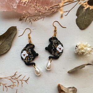 May include: Black teapot-shaped earrings with white floral details and gold accents. The earrings are dangling from gold hooks and have white pearl drops.