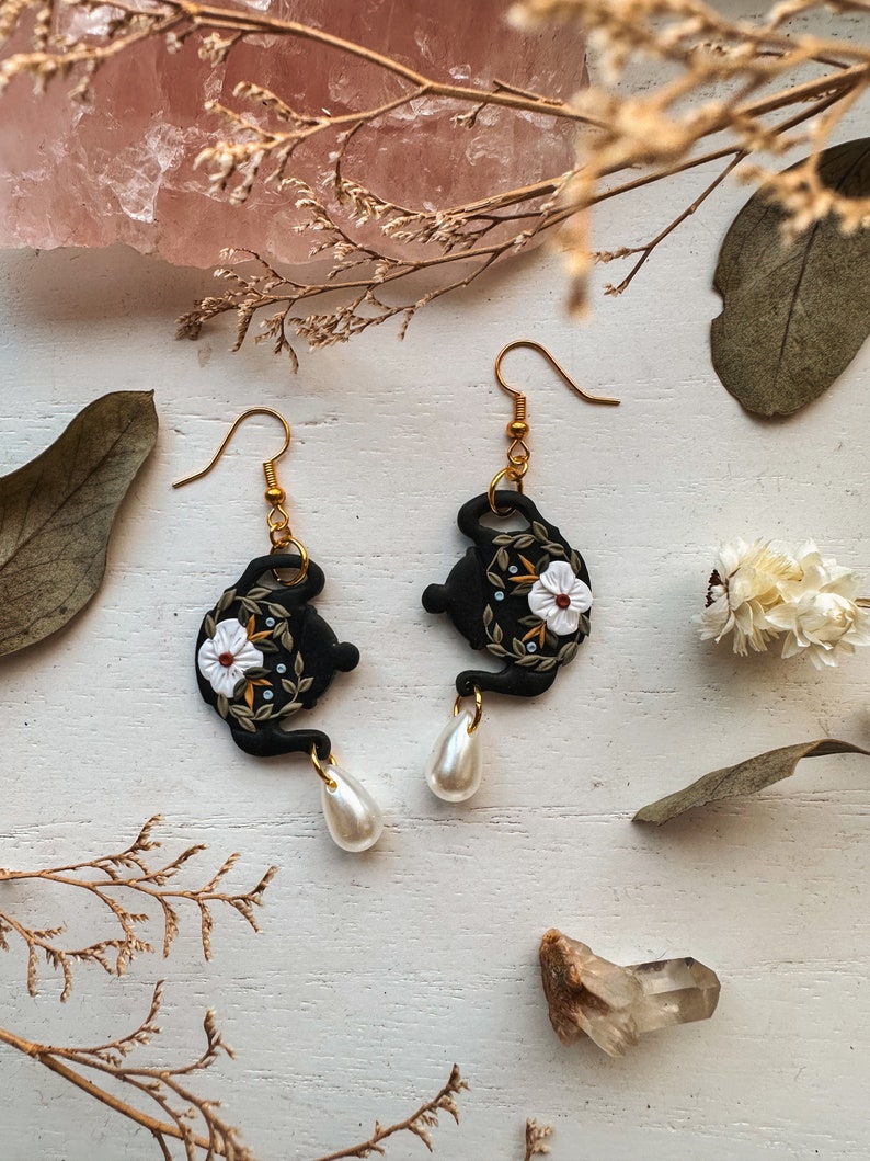 May include: Black teapot-shaped earrings with white floral details and gold accents. The earrings are dangling from gold hooks and have white pearl drops.