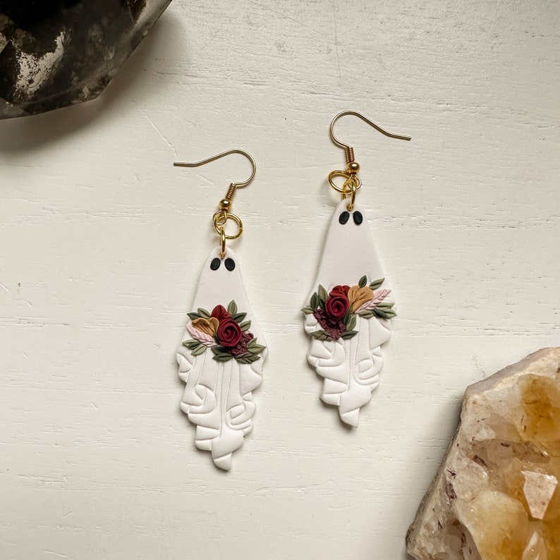May include: White ghost-shaped earrings with a floral design. The flowers are red, pink, and orange. The earrings are made of polymer clay and have gold hooks.