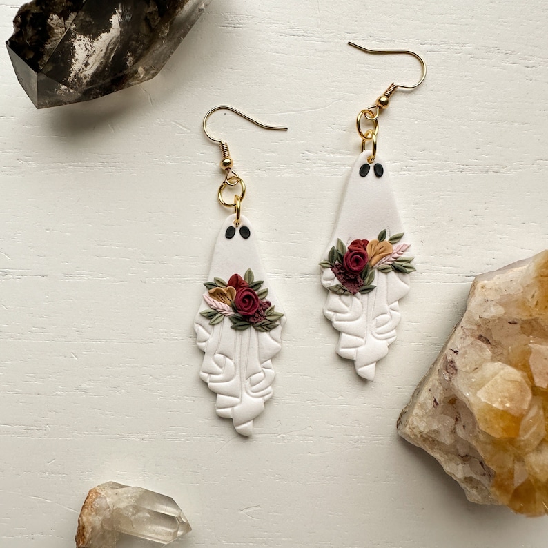May include: A pair of white ghost-shaped earrings with red and white floral accents. The earrings are made of polymer clay and have gold hooks.