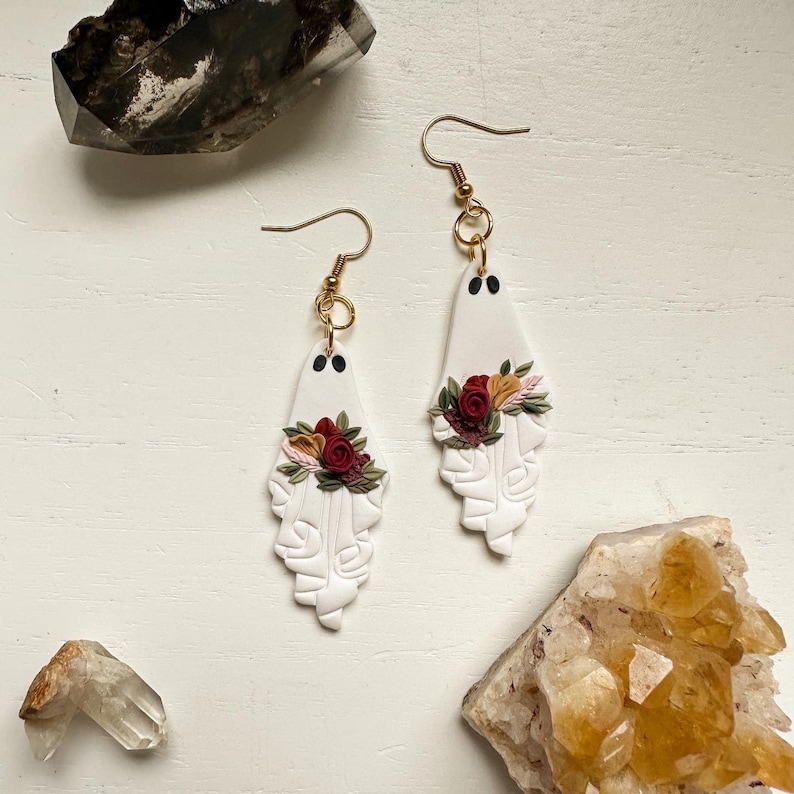 May include: A pair of white ghost-shaped earrings with red, pink, and yellow floral accents. The earrings are made of polymer clay and have gold hooks.