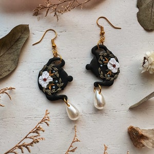 May include: Black teapot earrings with white floral details and a pearl dangle. The teapot is decorated with green leaves and a white flower with a yellow center.