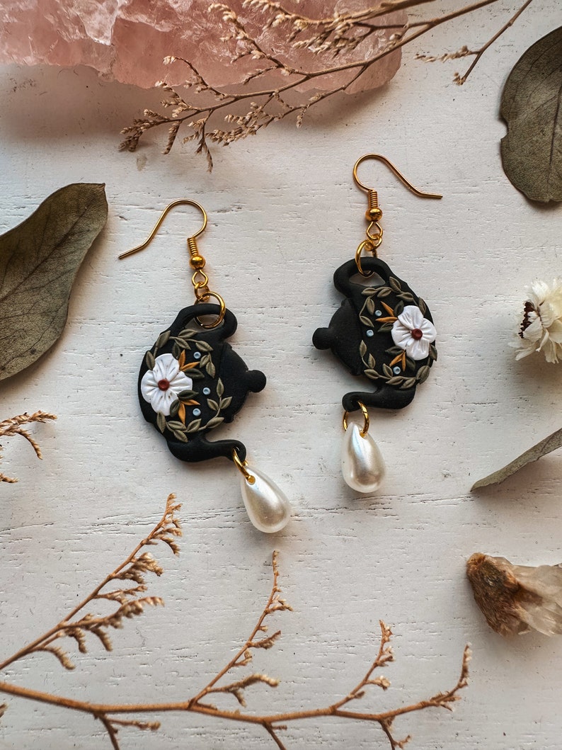 May include: Black teapot earrings with white floral details and a pearl dangle. The teapot is decorated with green leaves and a white flower with a yellow center.