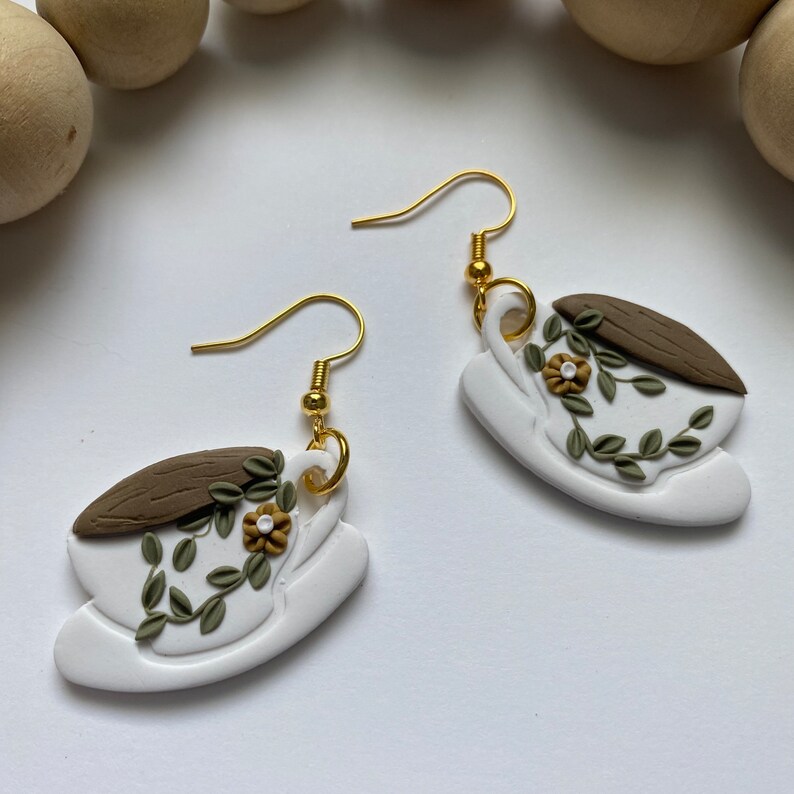 Puede incluir: Un par de pendientes de cer&aacute;mica blanca con forma de taza de t&eacute; con un borde marr&oacute;n y hojas verdes y un dise&ntilde;o de flores amarillas. Los pendientes tienen ganchos dorados.