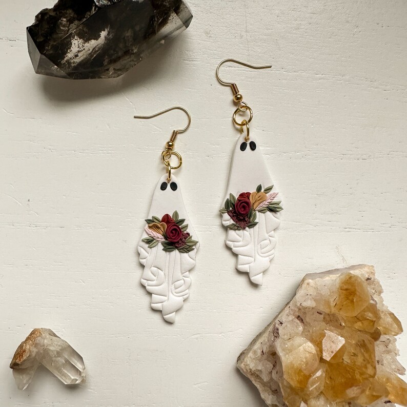 May include: A pair of white ghost-shaped earrings with red, white and green floral accents. The earrings are made of polymer clay and have gold hooks.
