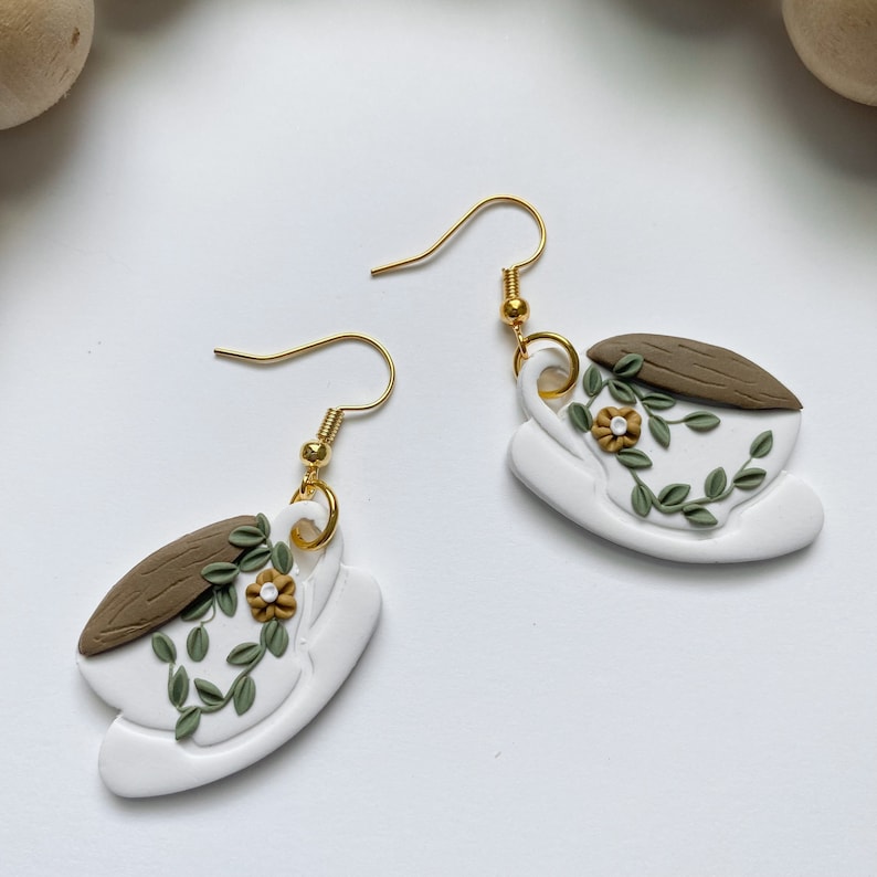 Puede incluir: Un par de pendientes de cer&aacute;mica blanca con forma de taza de t&eacute; con bordes marrones y hojas verdes y flores amarillas. Los pendientes tienen ganchos dorados.