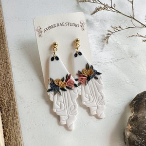 May include: A pair of white ceramic earrings with a floral design. The earrings are shaped like a teardrop and have a gold-colored hook. The flowers are in shades of pink, yellow, and brown.