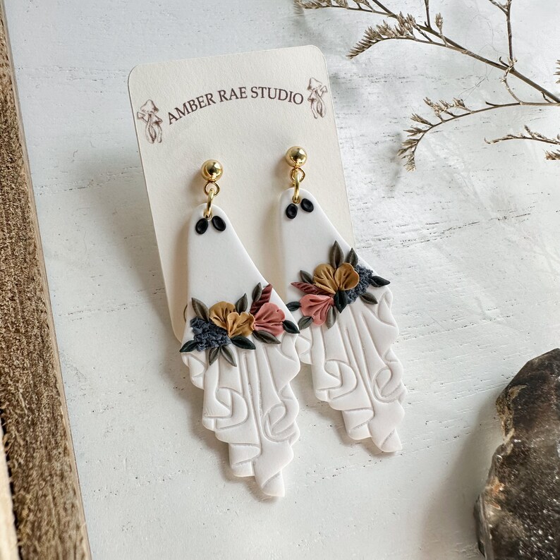 May include: A pair of white ceramic earrings with a floral design. The earrings are shaped like a teardrop and have a gold-colored hook. The flowers are in shades of pink, yellow, and brown.