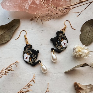 May include: Black teapot earrings with white floral details and gold accents. Each earring features a pearl dangle.