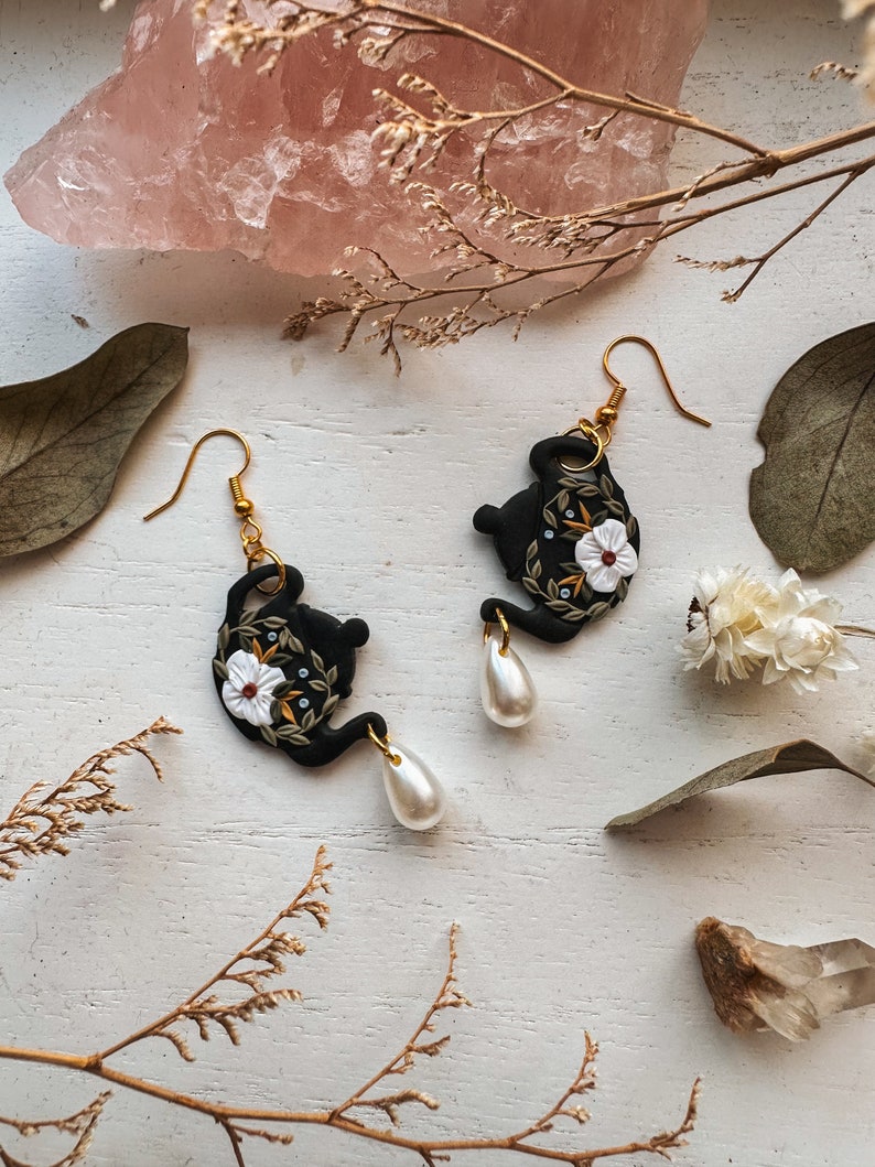 May include: Black teapot earrings with white floral details and gold accents. Each earring features a pearl dangle.