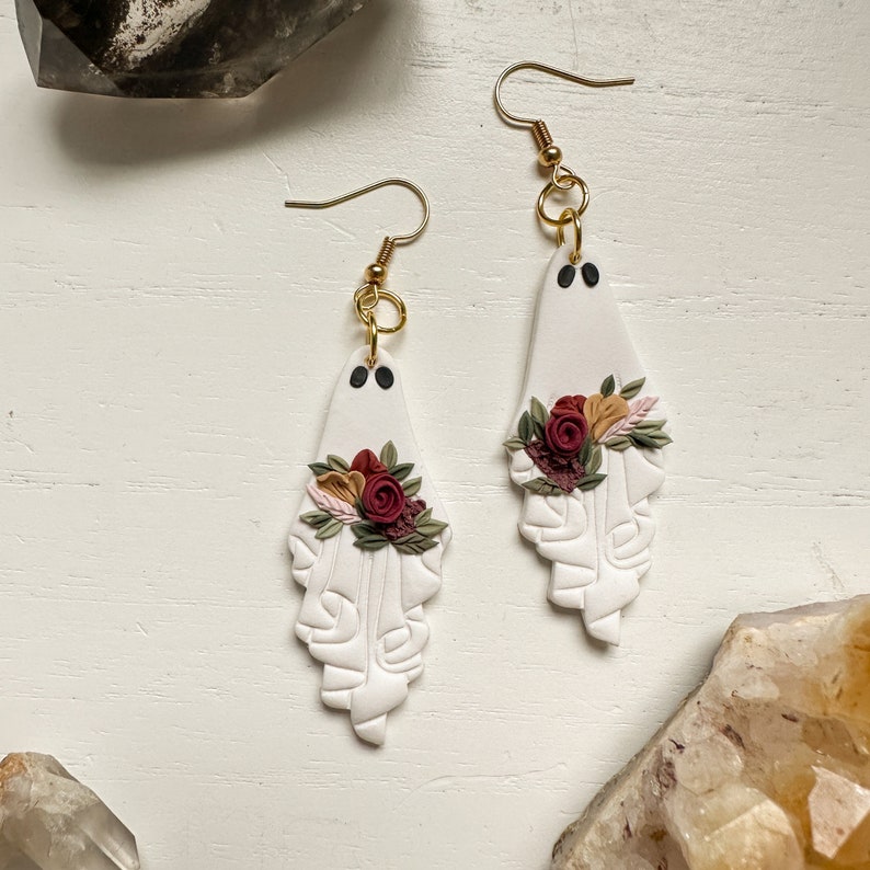 May include: A pair of white ghost-shaped earrings with red and pink floral accents. The earrings are made of polymer clay and have gold hooks.