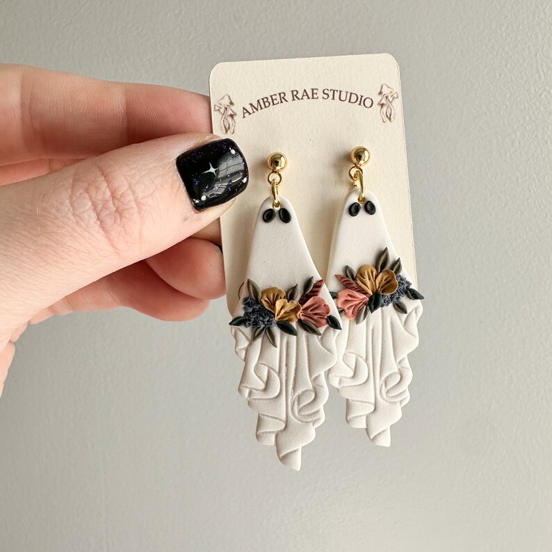May include: White ceramic earrings with a floral design in shades of pink, orange, and gold. The earrings are shaped like a draped fabric with a textured surface. The earrings are attached to gold hooks.