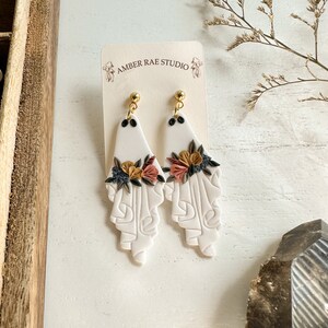 May include: White ceramic earrings with a floral design and gold accents. The earrings are shaped like a teardrop and have a textured surface. The flowers are in shades of pink, yellow, and brown. The earrings are hanging from gold hooks.
