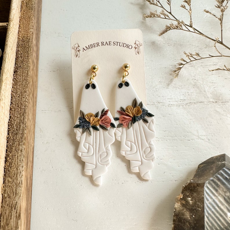 May include: White ceramic earrings with a floral design and gold accents. The earrings are shaped like a teardrop and have a textured surface. The flowers are in shades of pink, yellow, and brown. The earrings are hanging from gold hooks.