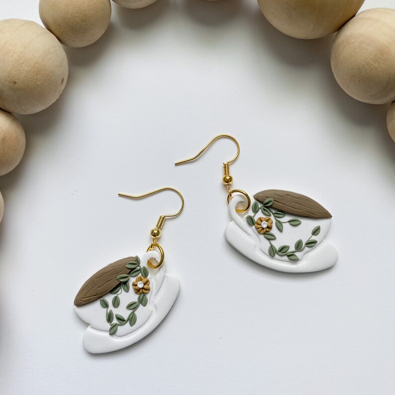 Puede incluir: Un par de pendientes de oro con tazas de t&eacute; de cer&aacute;mica blanca decoradas con hojas verdes y flores amarillas.