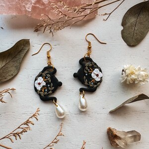 May include: A pair of black teapot earrings with white floral details and pearl drop accents.