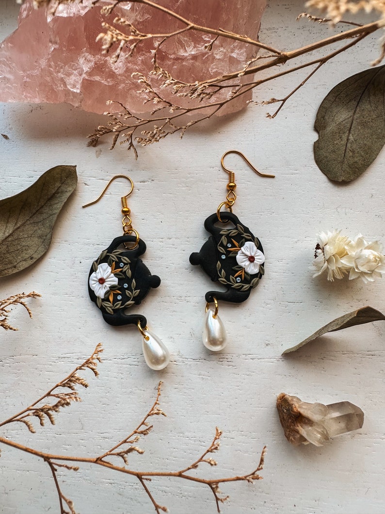 May include: A pair of black teapot earrings with white floral details and pearl drop accents.
