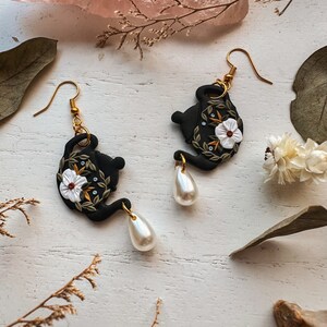 May include: A pair of black teapot earrings with white flowers and gold accents. Each earring features a pearl drop.