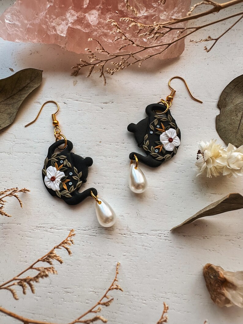 May include: A pair of black teapot earrings with white flowers and gold accents. Each earring features a pearl drop.