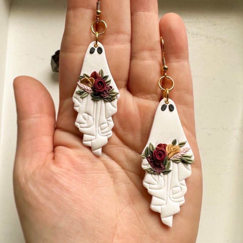 May include: A pair of white ghost-shaped earrings with burgundy and yellow floral accents. The earrings are made of polymer clay and have gold hooks.