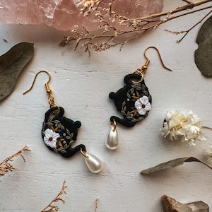 May include: A pair of black teapot earrings with white floral details and pearl accents. The earrings are on a white surface with dried flowers and a pink crystal.