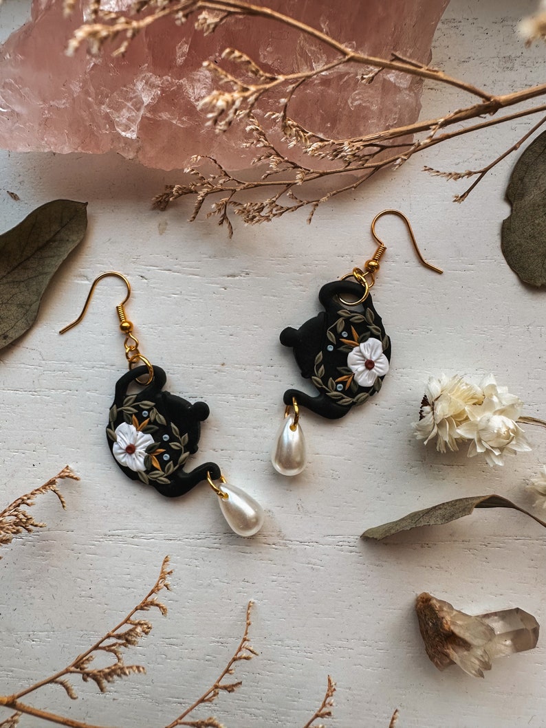 May include: A pair of black teapot earrings with white floral details and pearl accents. The earrings are on a white surface with dried flowers and a pink crystal.