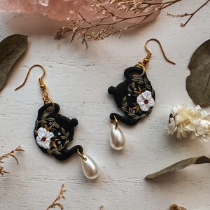 May include: A pair of black teapot earrings with white flowers and green leaves. Each earring has a dangling pearl.