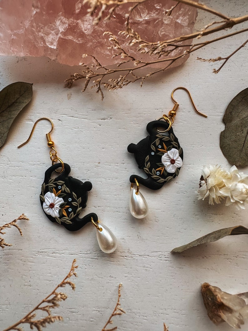May include: A pair of black teapot earrings with white flowers and green leaves. Each earring has a dangling pearl.
