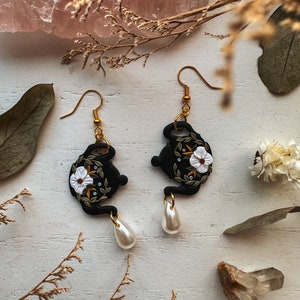 May include: Black teapot-shaped earrings with white floral details and gold accents. Each earring features a pearl drop.