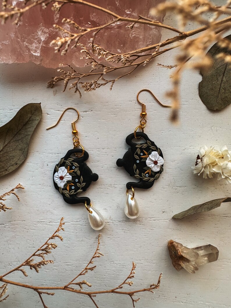 May include: Black teapot-shaped earrings with white floral details and gold accents. Each earring features a pearl drop.