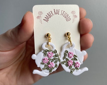 May include: White teapot-shaped earrings with pink flowers and green leaves. The earrings have gold hooks and are labeled "Amber Rae Studio".