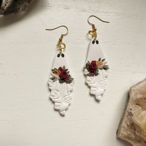 May include: White ghost-shaped earrings with a floral design. The flowers are red, pink, and yellow. The earrings have gold hooks.