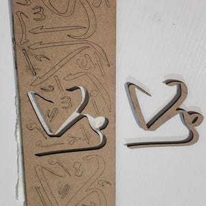 May include: A rectangular brown fibreboard sheet featuring laser-cut outlines of various Arabic calligraphy characters. One specific character shape has been removed from the centre of the board and is placed separately to the right of the sheet.