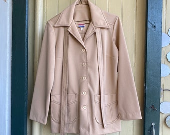 May include: A beige long-sleeve jacket featuring a wide collar, five front buttons, and two patch pockets. The garment includes two long, narrow fabric strips attached at the shoulders that hang down the front. A small label is visible at the neck.