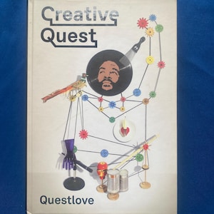 Könnte beinhalten: Ein gebundenes Buch mit dem Titel Creative Quest von Questlove mit weißem Einband. Das Cover zeigt eine Collage aus einem Gesicht auf einer Schallplatte, einem Vogel, bunten Zahnrädern, einem violetten Handschuh, Konservendosen und einer Kerze.