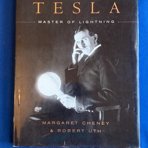 May include: A hardcover book titled Tesla: Master of Lightning by Margaret Cheney and Robert Uth. The black cover features a monochromatic portrait of Nikola Tesla holding a glowing, circular light bulb, with the title in gold lettering.