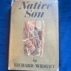 Può includere: Un libro con copertina rigida intitolato Native Son di Richard Wright. La sovraccoperta color marrone chiaro presenta un'illustrazione centrale di una persona in piedi su dei gradini vicino all'ingresso di un edificio, con un cestino vicino. Titolo e autore in nero.