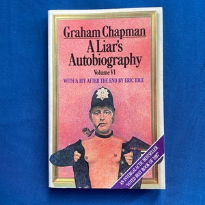 May include: A paperback book titled Graham Chapman A Liar's Autobiography Volume VI with a bit after the end by Eric Idle. The cover features an illustration of a person in a police helmet and pipe, opening a dark jacket against a pink background.