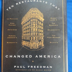 May include: A black hardback book titled Ten Restaurants That Changed America by Paul Freedman, with an introduction by Danny Meyer. The cover features a gold line drawing of a building and lists ten restaurants including Le Pavillon and Sylvia's.