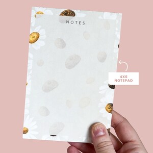 May include: A white notepad with a floral pattern of white daisies and brown centres. The notepad is labelled "NOTES" and says "4x6 NOTE PAD" on the side.