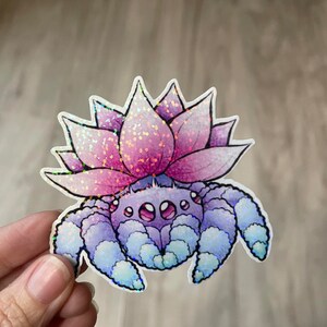 May include: A holographic sticker of a purple spider with a pink flower on its head. The spider has three pairs of legs and large eyes. The flower has a pink and purple gradient.