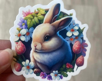 May include: A white rabbit with blue fur sits in a circle of colorful flowers. The flowers are pink, purple, blue, and white. The rabbit has pink ears and a pink nose.