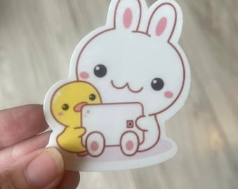May include: A white bunny with pink ears and a yellow duckling are holding a white phone with a pink camera lens. The bunny is wearing a pink bow and the duckling is wearing a pink bow. The bunny and duckling are sitting on a white background.