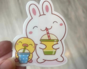 May include: A white bunny and a yellow chick are holding colorful cups with straws. The bunny is smiling and has pink ears. The chick is also smiling and has a small beak. The background is a light brown wood.