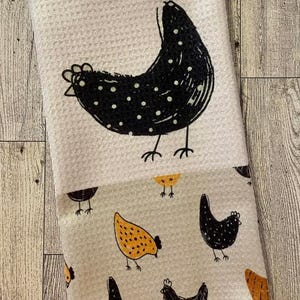 May include: White and gray kitchen towel with a repeating chicken pattern. The top section features a large black chicken with white polka dots. The bottom section has smaller chicken illustrations in black and yellow.