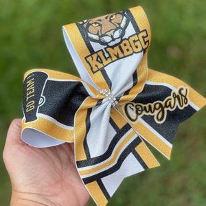 May include: A white, black, and gold cheer bow with a cougar mascot, the text "Go Team!" and "KLM BGC Cougars".