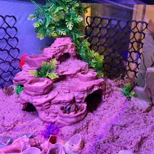 May include: A pink, multi-level rock-like structure for a terrarium, featuring several circular basins, hollow tunnels, and artificial green foliage. A black heart-patterned mesh divider stands behind the structure on a sandy substrate.
