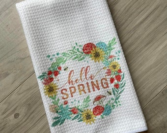 May include: White kitchen towel with a floral wreath design and the text "hello spring" in orange. The wreath includes sunflowers, red roses, berries, and mushrooms.