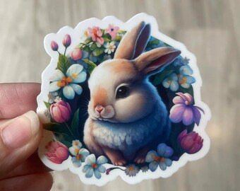 May include: A white rabbit with blue fur sits in a circle of colorful flowers. The rabbit has pink ears and a pink nose. The flowers are pink, blue, and purple.