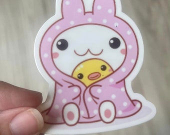 May include: A pink bunny wearing a pink and white polka dot hooded robe with a yellow duckling inside.